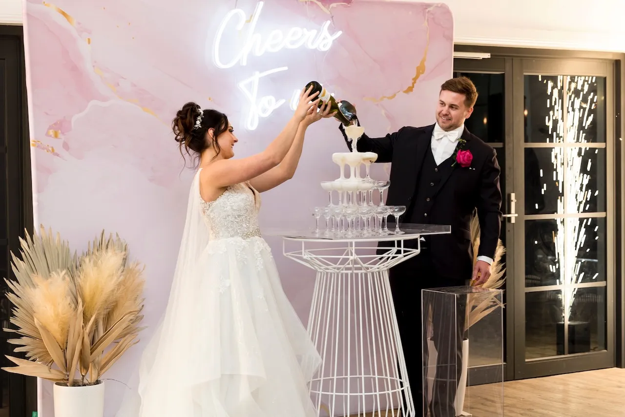 Photo of bride and groom pouring sparkling wine onto tower of champagne glasses